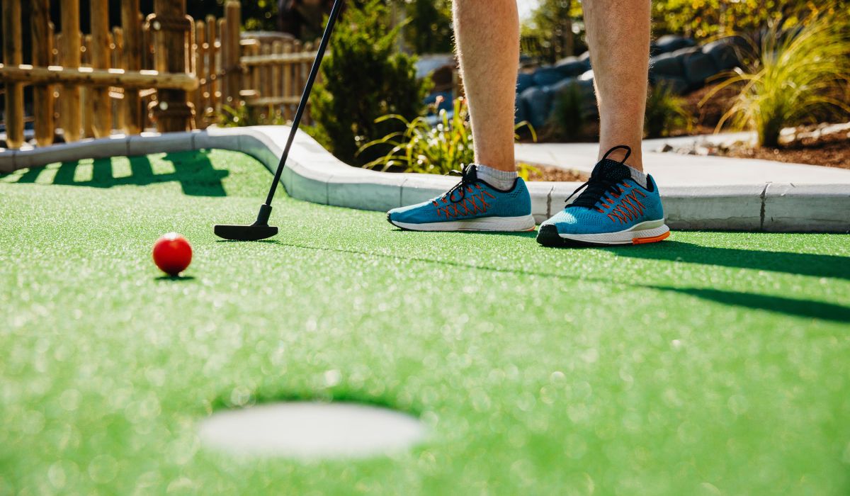 Magnolia Person in blue sneakers playing mini-golf, holding a putter near a red ball on a green course, aiming for the hole.